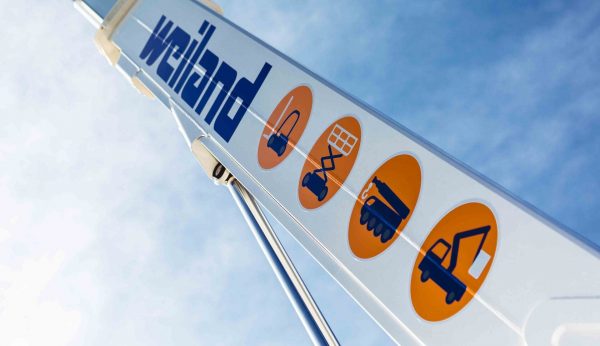 Detailaufnahme eines Weiland-Kranarms mit Piktogrammen für verschiedene Hebe- und Transportgeräte vor blauem Himmel.